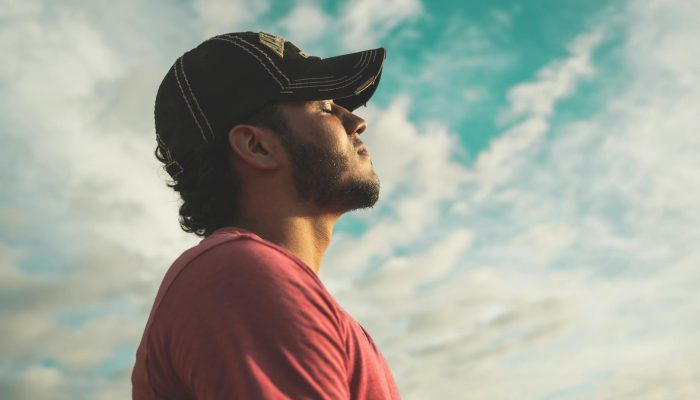 man wearing black cap with eyes closed under cloudy sky